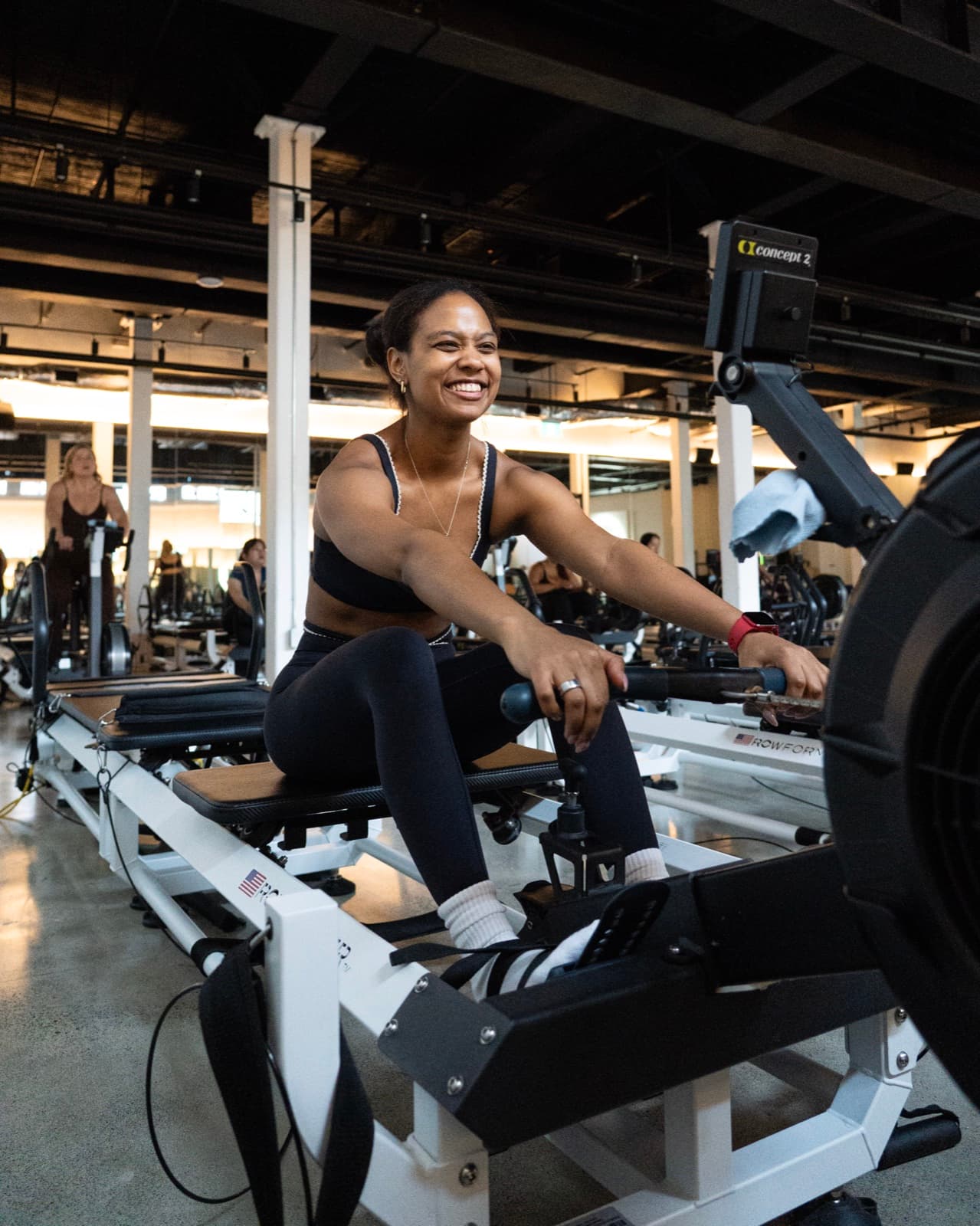 A woman smiling mid-stroke on a rowing machine in a large gym