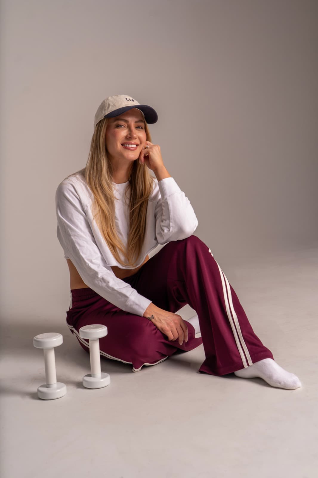 A smiling woman in a cap and burgundy track pants seated on the studio floor beside white dumbbells