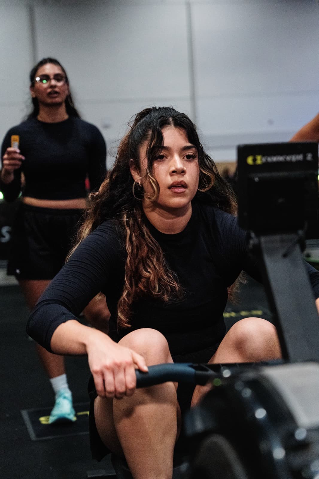 A rower mid-stroke on a Concept2, her partner watching behind her