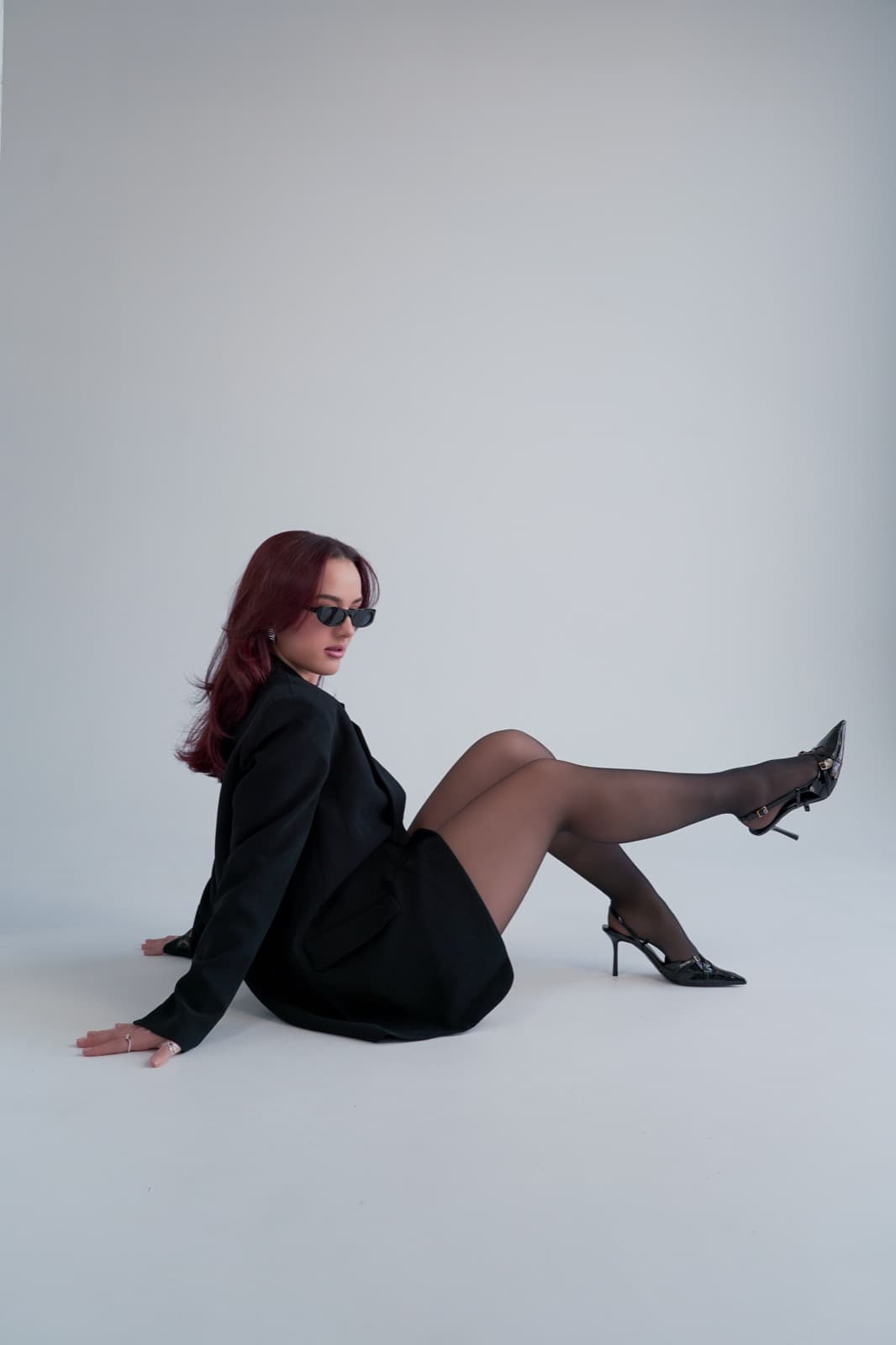Seated on the studio floor in sunglasses, a black blazer and sheer tights, one leg raised