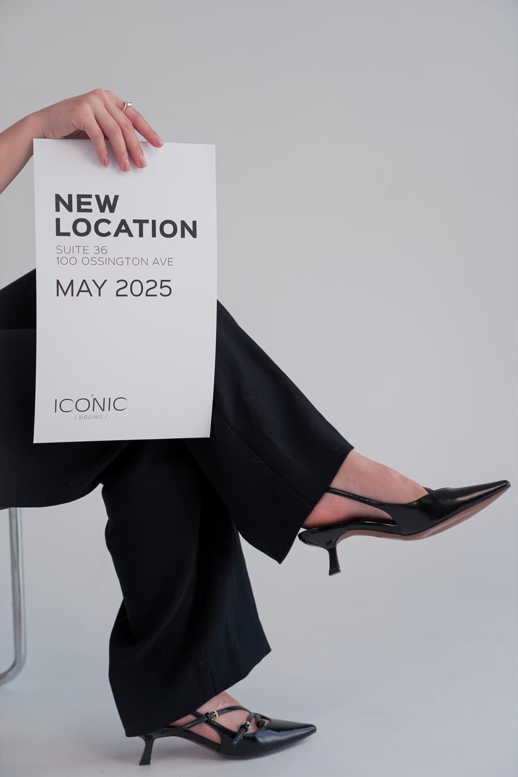 Crossed legs in black trousers and kitten heels behind a New Location, May 2025 sign